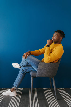 Relaxed young African man in casual wear sitting on the chair against the blue wallの写真素材
