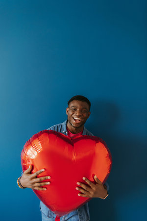 Joyful young African man holding heart shaped balloon and smiling while standing on red background