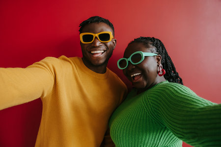 Fashionable young African couple in trendy glasses embracing and smiling on red background togetherの写真素材