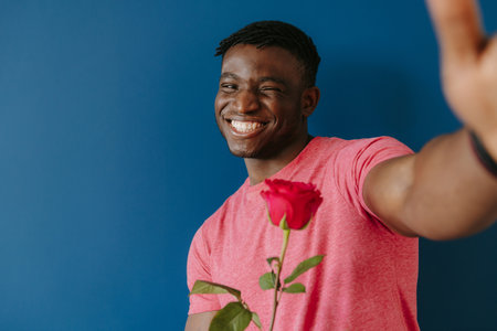 Happy young African man in casual wear making selfie while holding single rose on blue backgroundの写真素材