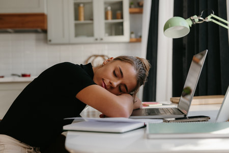 Tired young businesswoman leaning head on laptop and sleeping while sitting at the desk at homeの写真素材