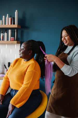 Confident African American hairdresser braiding hair to a smiling female customer in salonの写真素材