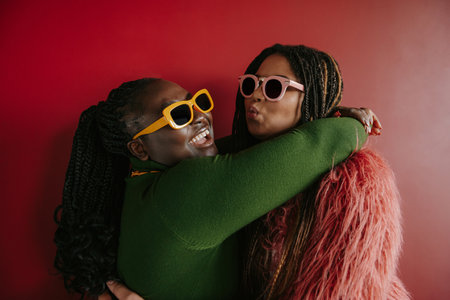 Two joyful plus size African woman with braided hair embracing on red background togetherの写真素材
