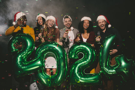 Joyful people in Christmas hats holding 2024 numbers while celebrating New Year in night clubの写真素材