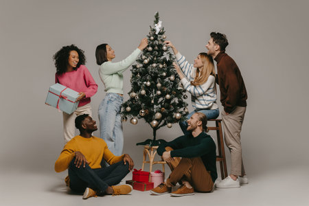 Group of joyful young people having fun while decorating Christmas tree on studio background
