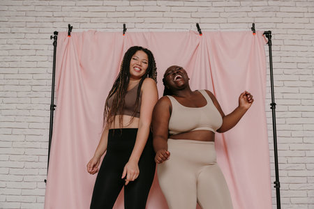 Two happy plus size African women in sportswear dancing while standing on fabric background togetherの写真素材
