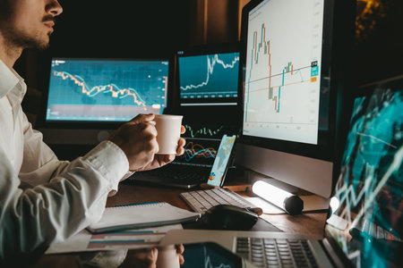 Male trader holding coffee cup while analyzing stock market data on multiple computers at office