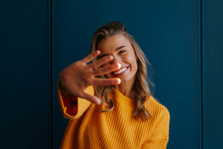Shy young blond hair woman in yellow sweater stretching out hand on blue backgroundの写真素材