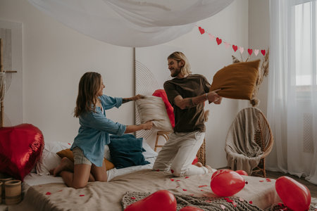Smiling young loving couple having a pillow fight in bed surrounded with red heart shape balloonsの写真素材