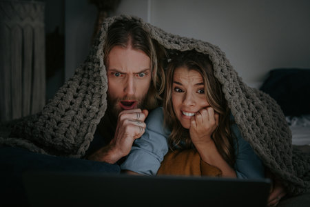 Terrified young couple covering with blanket and watching horror movie on laptop while lying in bedの写真素材