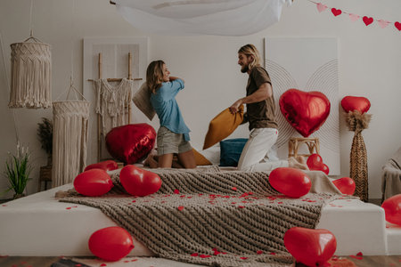 Happy young loving couple having a pillow fight in bed surrounded with red heart shape balloonsの写真素材