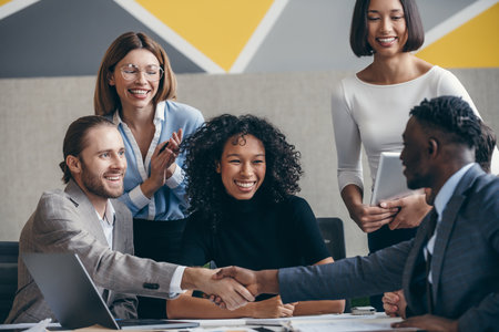 Two happy young businessmen shaking hands while sitting with their colleagues at the office deskの写真素材