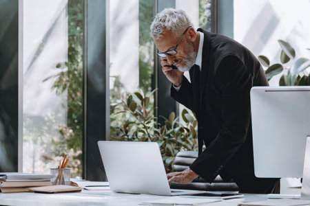 Concentrated mature businessman talking on mobile phone and using computer while working in officeの写真素材