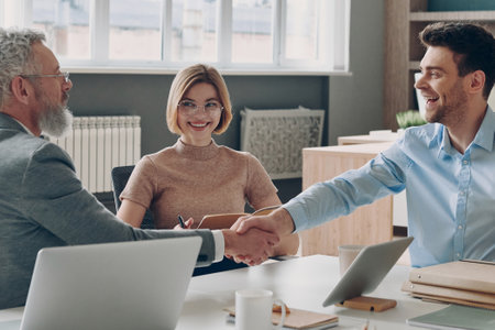 Cheerful business people shaking hands while sitting at the office desk togetherの写真素材