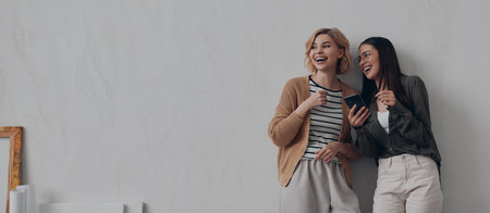 Two cheerful young women talking and smiling while standing near their creative working placeの写真素材