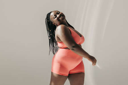 Happy African plus size woman in sportswear radiating self love while standing in fitness studio