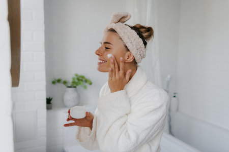 Happy young woman in bathrobe applying face cream while standing in bathroomの写真素材