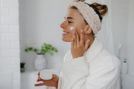 Happy young woman in bathrobe applying face cream while standing in bathroomの写真素材