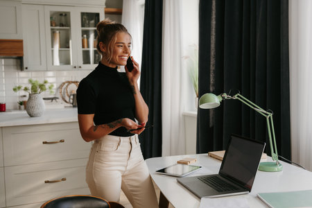 Confident young woman talking on mobile phone and smiling while working at homeの写真素材