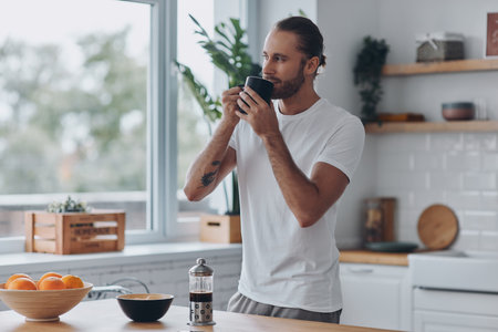 Dreamful young man enjoying fresh coffee while standing at the kitchenの写真素材
