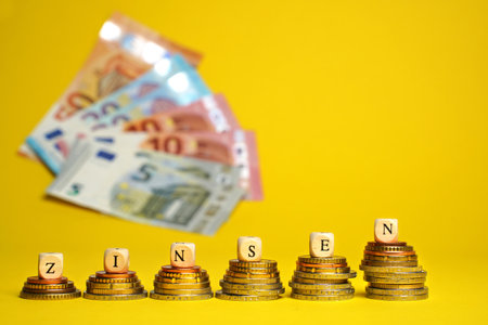 German word for interest charges, ZINSEN, from wood cubes on stacks of coins. Euro cash money on yellow background, coins and banknotes, close up.の写真素材
