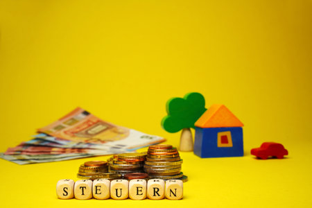 German word for tax, Steuern, from wood cubes with letter. Euro cash money, wood house, tree and auto on yellow background, stacks of coins, banknotes, close up.の写真素材
