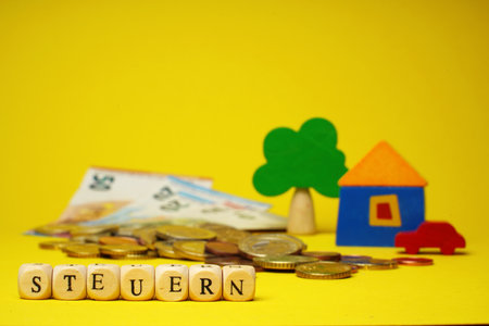German word for tax, Steuern, from wood cubes with letter. Euro cash money, wood toy house, tree and auto on yellow background, euro coins and banknotes, close up, copy space.の写真素材
