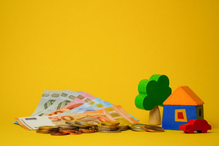 Euro cash money euro coins and banknotes with wood toy house, tree and auto on yellow background. Property Ownership Concept.の写真素材