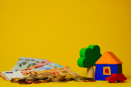 Euro cash money euro coins and banknotes with wood toy house, tree and auto on yellow background. Property Ownership Concept.の写真素材