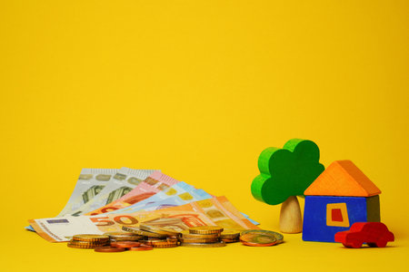 Euro cash money euro coins and banknotes with wood toy house, tree and auto on yellow background. Property Ownership Concept.の写真素材