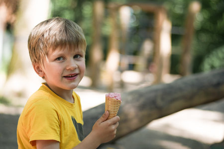 Boy kid in yellow t-shirt holding strawberry ice cream in waffles cone outdoor. summer time at the playground. copy spaceの写真素材