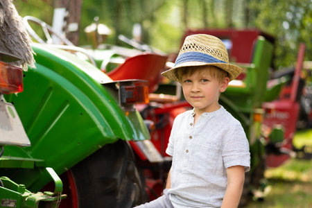 Happy cute little boy in a hat standing next to a red and green tractors.の写真素材
