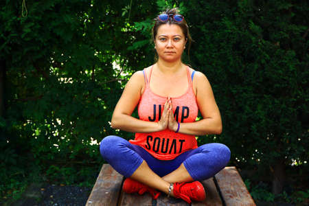 Yoga instructor shows sukhasana, easy pose on wooden bench at back yard.の写真素材
