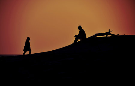 Silhouettes of a woman walking towards a man that is sitting at a beach during  sunset