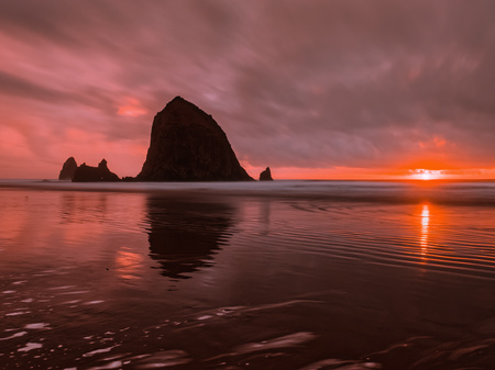 Haystack Rock silhouette reflects in the water at Cannon Beach, Oregon. Flaming red afterglow as sun sets on the horizon, long exposure shot with clouds in motion over Pacific Ocean.の写真素材
