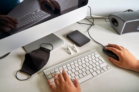 A child's hands on a computer keyboard next to a mask by the covid-19 in his bedroom. Concept to...
