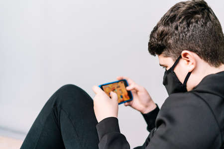 A young teenager wearing a mask plays with a smartphone online sitting on the street.の写真素材