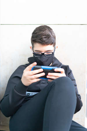 A young teenager wearing a mask plays with a smartphone online sitting on the street.の写真素材