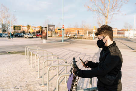Electric scooter and cell phone. Masked teenager by  uses electronic vehicle rental service with smartphone in urban city street and park. Boy uses cell phone for eco-friendly transportation rental.の写真素材