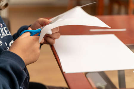 detail of a girl's hands cutting with a pair of children's scissors some sheets of paper to make crafts at home.の写真素材