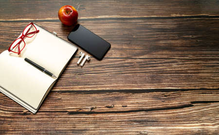 Open notebook with empty pages and other office supplies, glasses a smartphone and a red apple on a wooden desk table. Top view, plan and copy spaceの写真素材