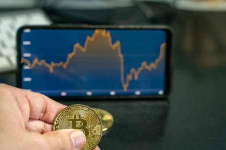 a hand holding a bitcoin coin with a smartphone depicting graphs of the cryptocurrency market. Digital currency concept.の写真素材