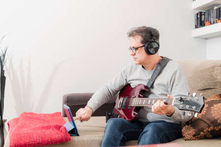 50-year-old concentrated man follows teacher's instructions via tablet to learn to play electric guitar at homeの写真素材