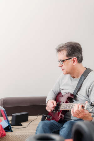 50-year-old concentrated man follows teacher's instructions via tablet to learn to play electric guitar at homeの写真素材