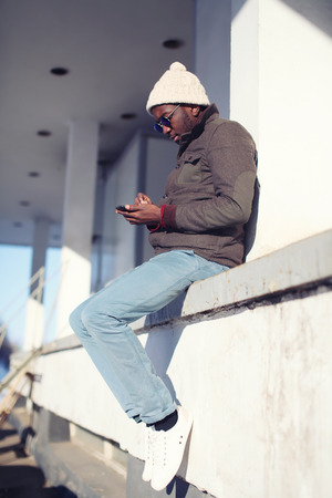 Street fashion and technology concept - lifestyle stylish young african man using smartphone in the cityの写真素材