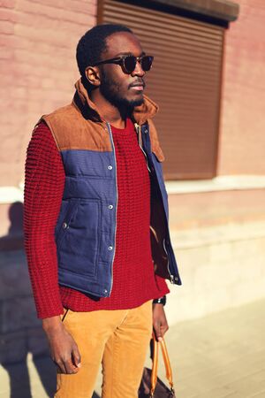 Fashion portrait of stylish confident young african man wearing a sunglasses and sweater with vest jacket evening in the cityの写真素材