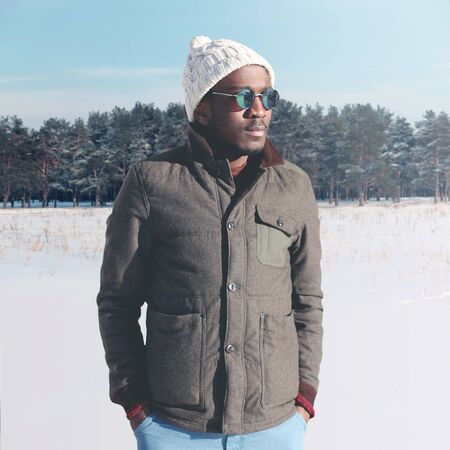 Fashion stylish young african man wearing a sunglasses and jacket with knitted hat in winter day over snowの写真素材