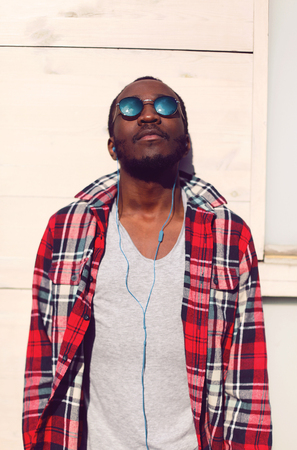 Fashion portrait of stylish young african man listens to music in the city, wearing a hipster plaid red shirt and sunglasses with earphonesの写真素材