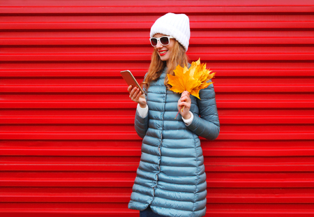 Fashion autumn smiling woman using smartphone holds yellow maple leaves in hands on a red background in the cityの写真素材
