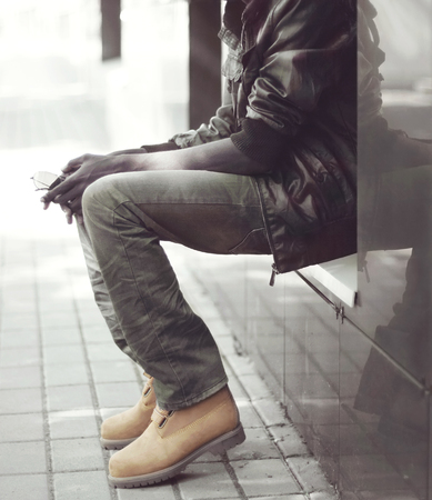 Fashion african man in black jacket, jeans and boots sitting in the cityの写真素材